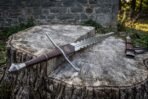 Wavy bladed sword with faceted pommel and brown wrapped grip laid diagonally across a cut stump by a stone wall