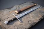 Wavy bladed sword with knotwork etched ricasso and black grip lying on slate beside a timber scabbard with leather band
