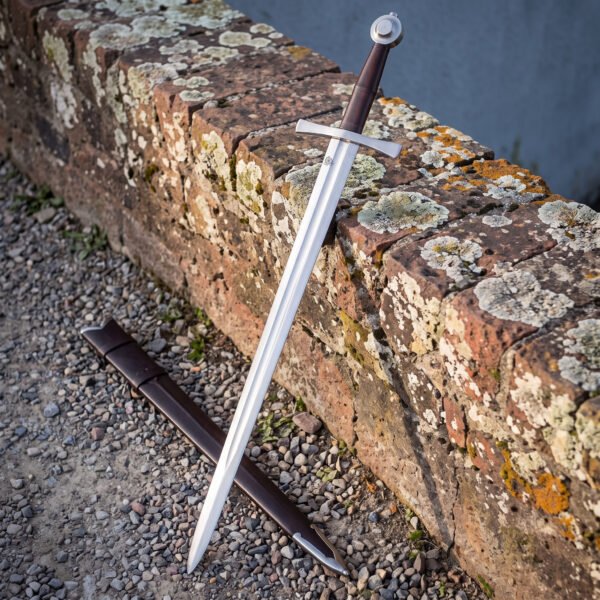 Wheel pommel knightly sword in 1095 carbon steel standing on a lichen covered wall, showing the dark brown leather grip, flared cross guard and brown leather scabbard