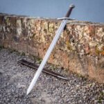 Wheel pommel knightly sword leaning against a lichen covered sandstone wall with its brown leather scabbard on the gravel below, blade polished and tapering to a point