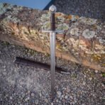 Wheel pommel knightly sword shown upright from the front with its brown leather scabbard on gravel, the long tapering polished blade and steel cross guard clearly visible