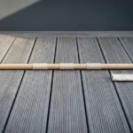 White ash bo staff with polished steel end ferrules and three whipped cord grip bands lying across grey timber decking beside its canvas carry sleeve