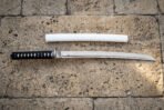 White lacquered saya wakizashi and its clay quenched 1095 steel blade laid flat above one another on pale limestone paving seen straight down in warm daylight