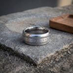 Wide silver tone Norse rune ring with an Elder Futhark centre band flanked by twisted rope borders standing on a grey stone slab beside a shallow wooden tray