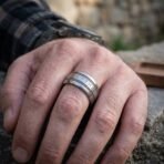 Wide silver tone rune ring with rope edged borders and rune band worn on a man's finger, plaid shirt cuff and stone wall behind