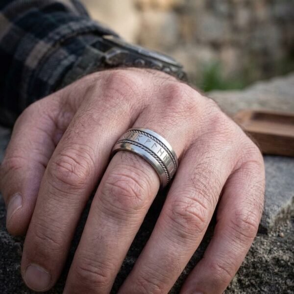 Wide silver tone rune ring with rope edged borders and rune band worn on a man's finger, plaid shirt cuff and stone wall behind