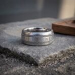 Wide silver tone rune ring with twisted rope borders and an elder futhark centre band standing on a stone slab, oak tray behind
