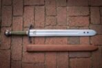Wolf etched arming sword in polished high carbon steel with a blackened curved guard, olive green leather grip, faceted pommel and leather scabbard on red brick paving