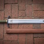 Wolf etched arming sword in polished high carbon steel with a blackened curved guard, olive green leather grip, faceted pommel and leather scabbard on red brick paving