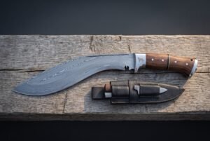 Wolf etched kukri knife in pattern welded damascus with rosewood scales lying on weathered grey planks beside its black leather sheath and two companion blades.