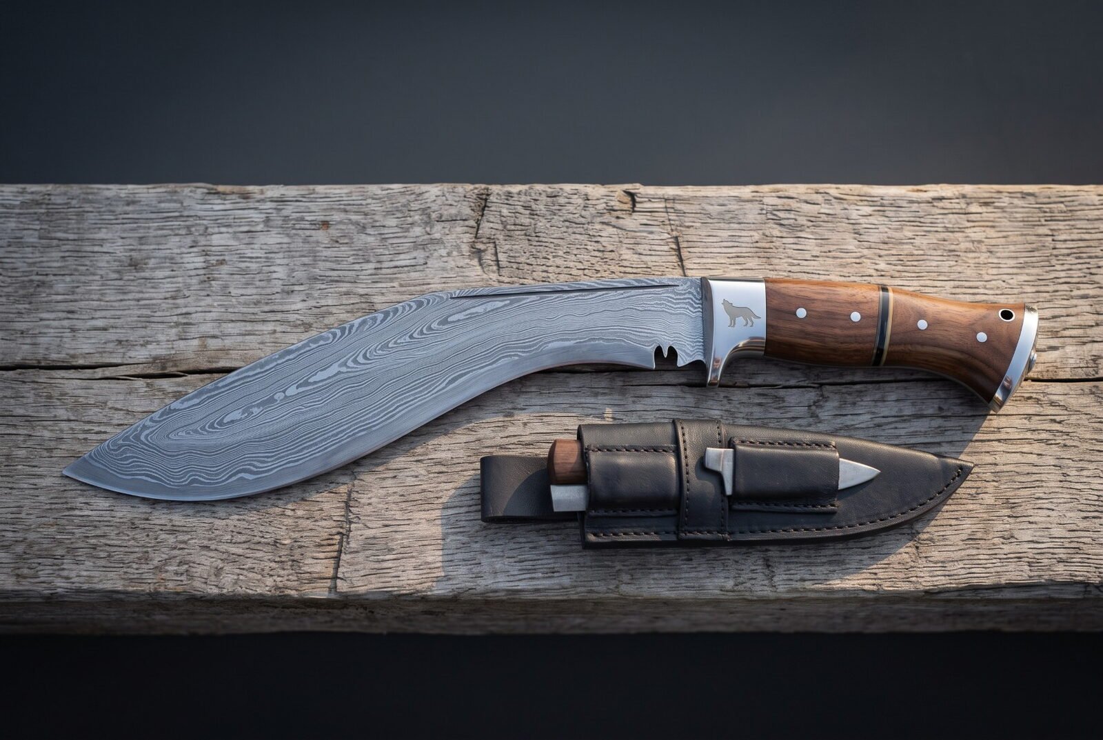 Wolf etched kukri knife in pattern welded damascus with rosewood scales lying on weathered grey planks beside its black Wolf etched kukri knife in pattern welded damascus with rosewood scales lying on weathered grey planks beside its black leather sheath and two companion blades.