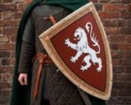 Wooden heater shield with a painted white rampant lion on a red field and carved knotwork border carried by a man in a green cloak
