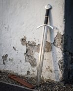 Yggdrasil arming sword with a pattern welded damascus blade leaning on a peeling whitewashed wall above gravel, beside its brown leather scabbard and brass banded grip.