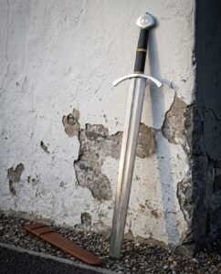 Yggdrasil arming sword with a pattern welded damascus blade leaning on a peeling whitewashed wall above gravel, beside its brown leather scabbard and brass banded grip.