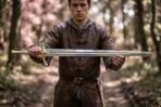 Young man in a leather jerkin holds a steel fantasy arming sword level across both hands on a woodland path